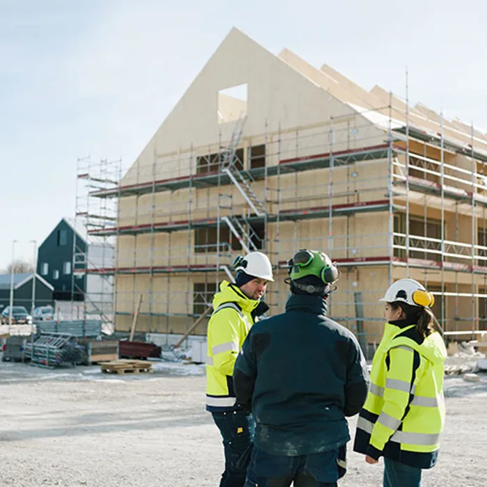 Tre personer står och pratar framför ett bygge där ett hus uppförs
