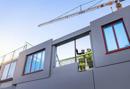 Två byggnadsarbetare står och tittar ut från ett bygge av ett hus.