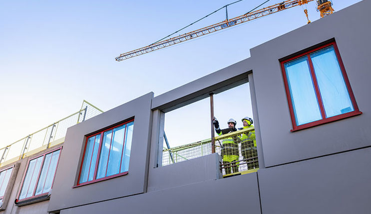 Två byggnadsarbetare står och tittar ut från ett bygge av ett hus.