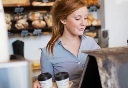 A girl stands behind the cashier counter holding two cups of coffee.