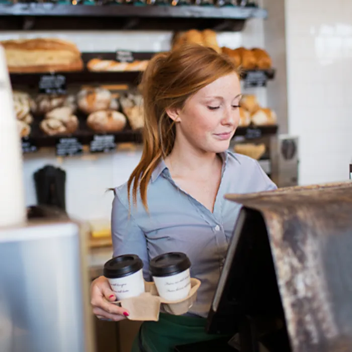 A girl stands behind the cashier counter holding two cups of coffee.
